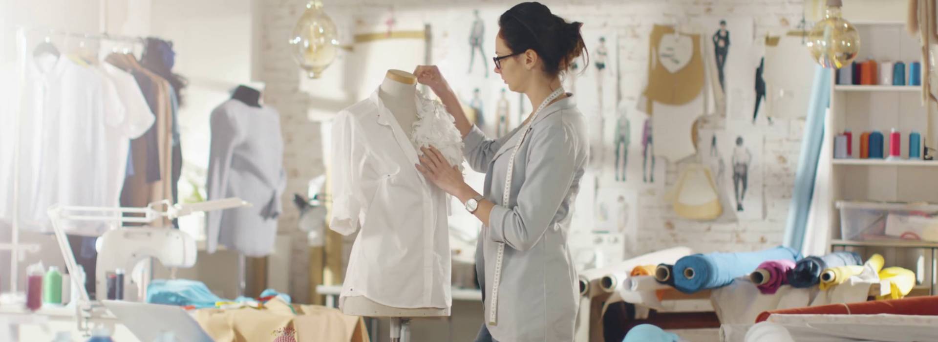 A studio tailor with a tape measure around her neck shaping a white shirt on a dress form, surrounded by rolls of fabric and spools of thread