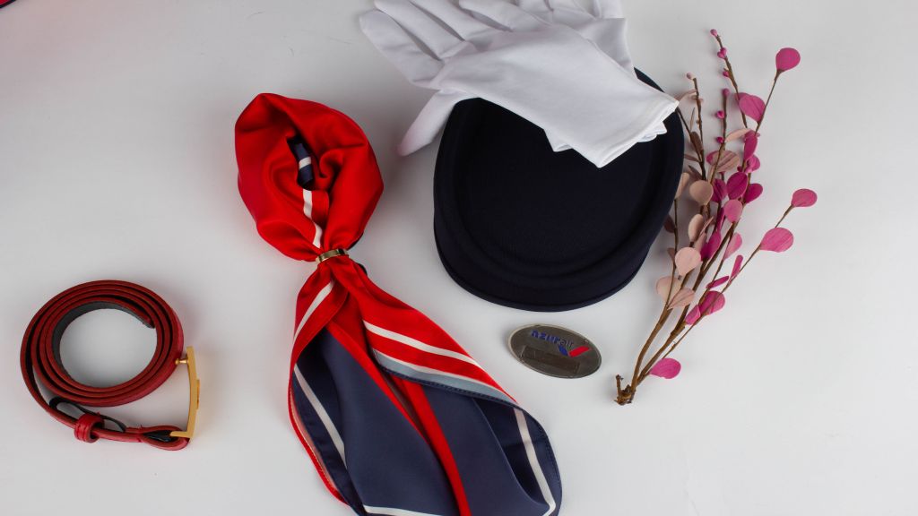 A flat lay of cabin-crew accessories, with a navy and red flight bag, a printed neck scarf, a pillbox hat, white gloves, a belt, and a blossom branch