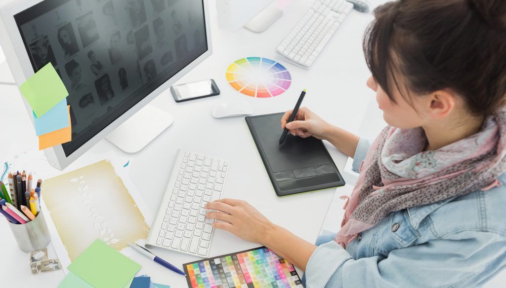 A designer working at a drawing tablet with a colour wheel and a fabric swatch fan on the desk beside the keyboard